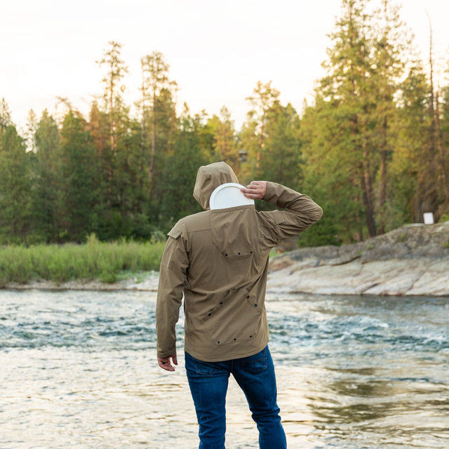 A person wearing a tan ODIN Disc Golf jacket with a hood, showcasing the back top pocket designed for disc storage.